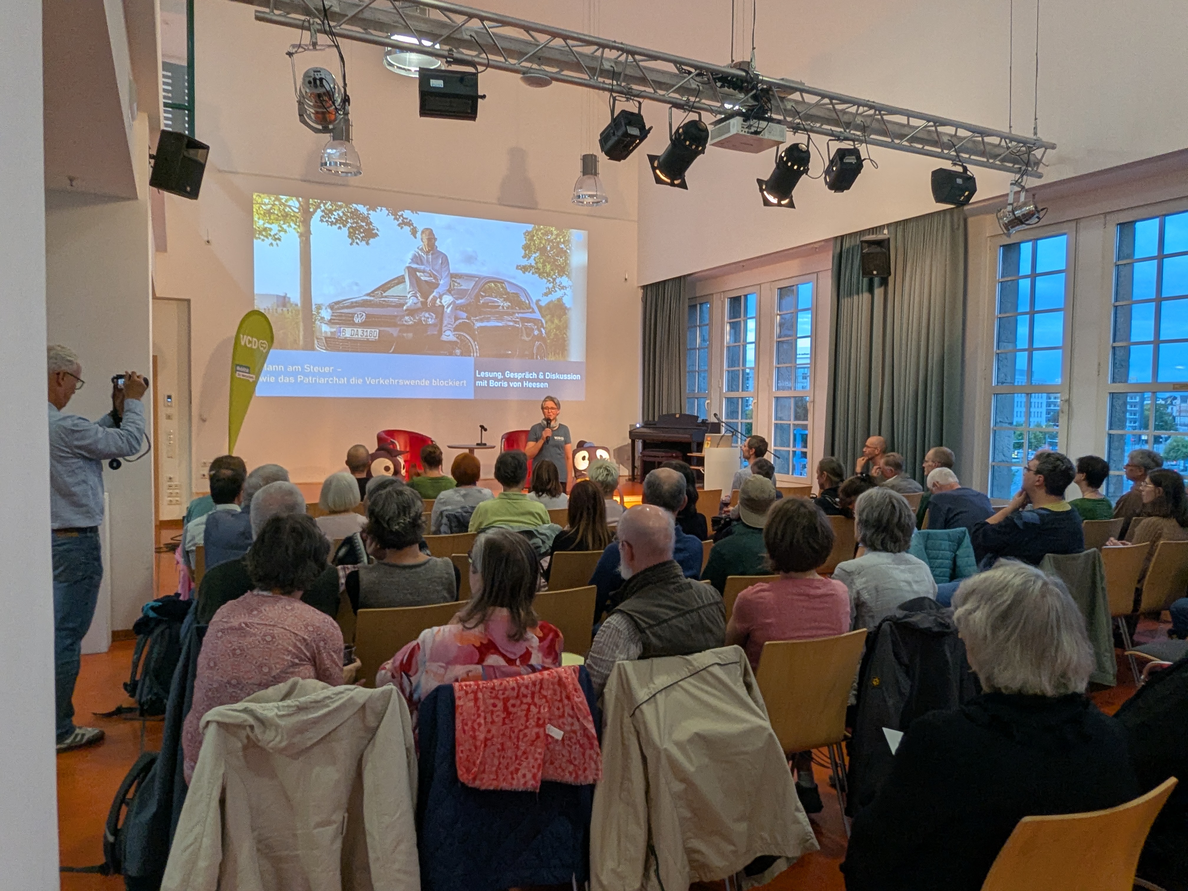 Vortrag mit Publikum in einem Veranstaltungssaal; auf der Leinwand ist die Präsentation „Mann am Steuer – wie das Patriarchat die Verkehrswende blockiert“ zu sehen.
